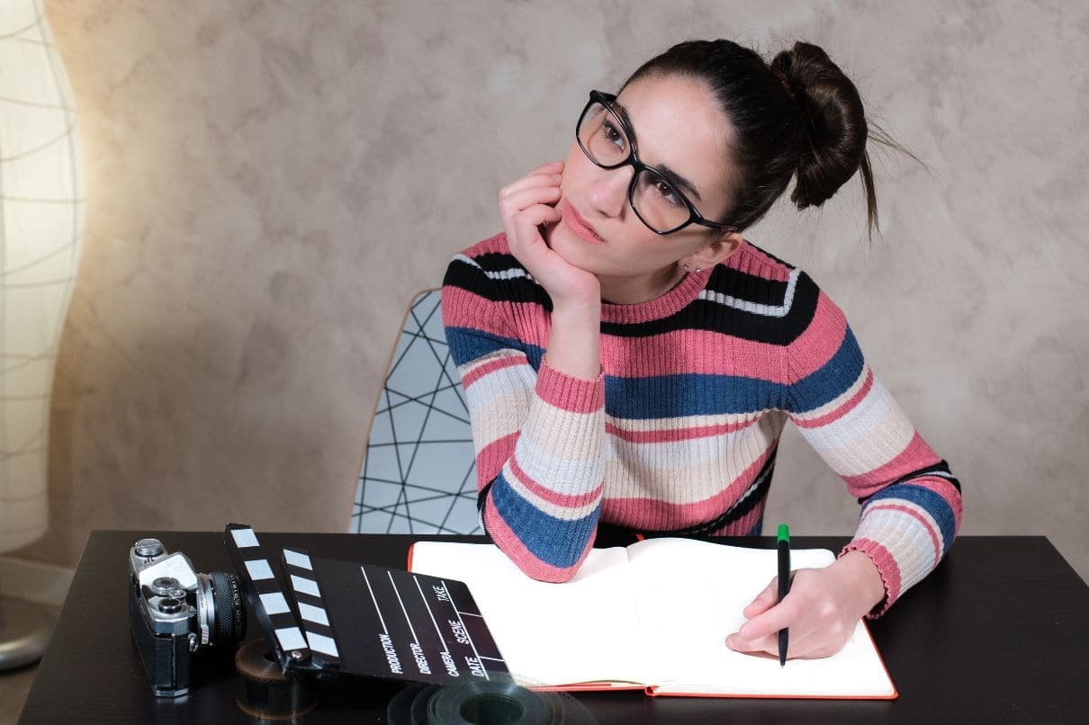 A woman with glasses sits at a desk, holding a notebook and camera, ready to capture her thoughts and ideas.